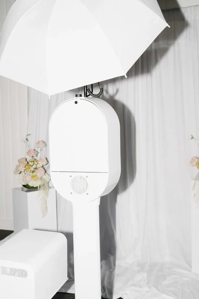 White open-air photo booth with a softbox light beside a white drapery backdrop and pale floral arrangements.