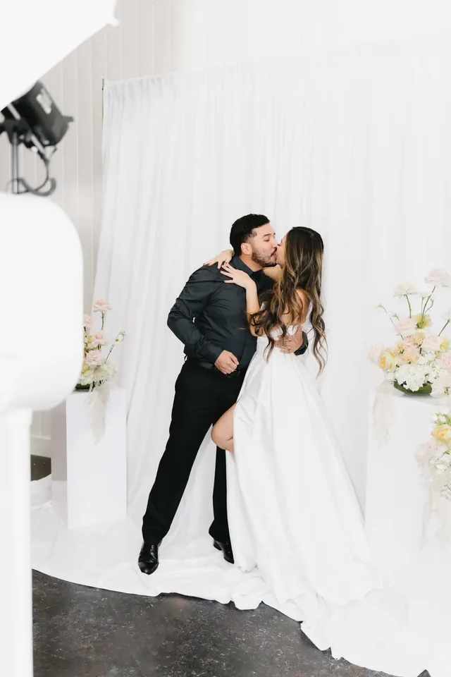 Couple in formalwear embracing in front of a white drapery backdrop with soft floral arrangements.